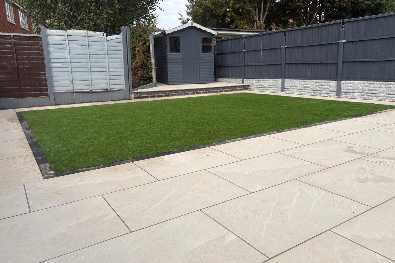 Finished garden with a large porcelain patio, artificial lawn edged in setts, painted fencing and a grey shed by AGG Renovations
