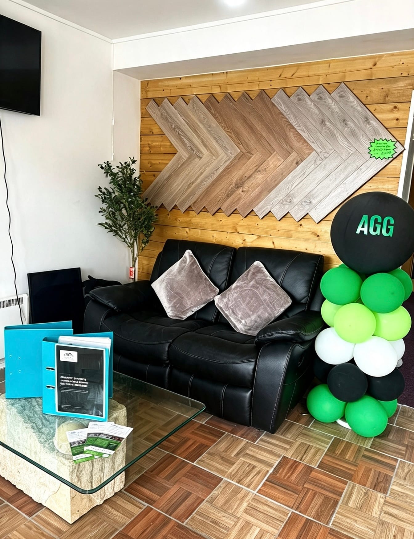 The seating area at AGG Renovations: a black leather sofa against a timber wall laid with a herringbone flooring display, a glass table with brochures, and green and black balloons